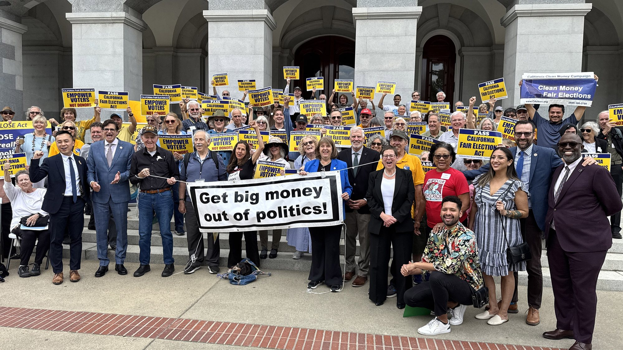Supporters and legislators on the Capitol steps holding a banner reading Get big money out of politics